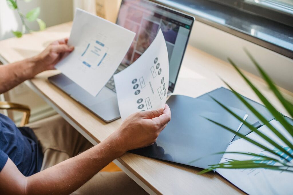 Crop anonymous male designer in casual wear sitting at table with opened laptop and folders and reading papers